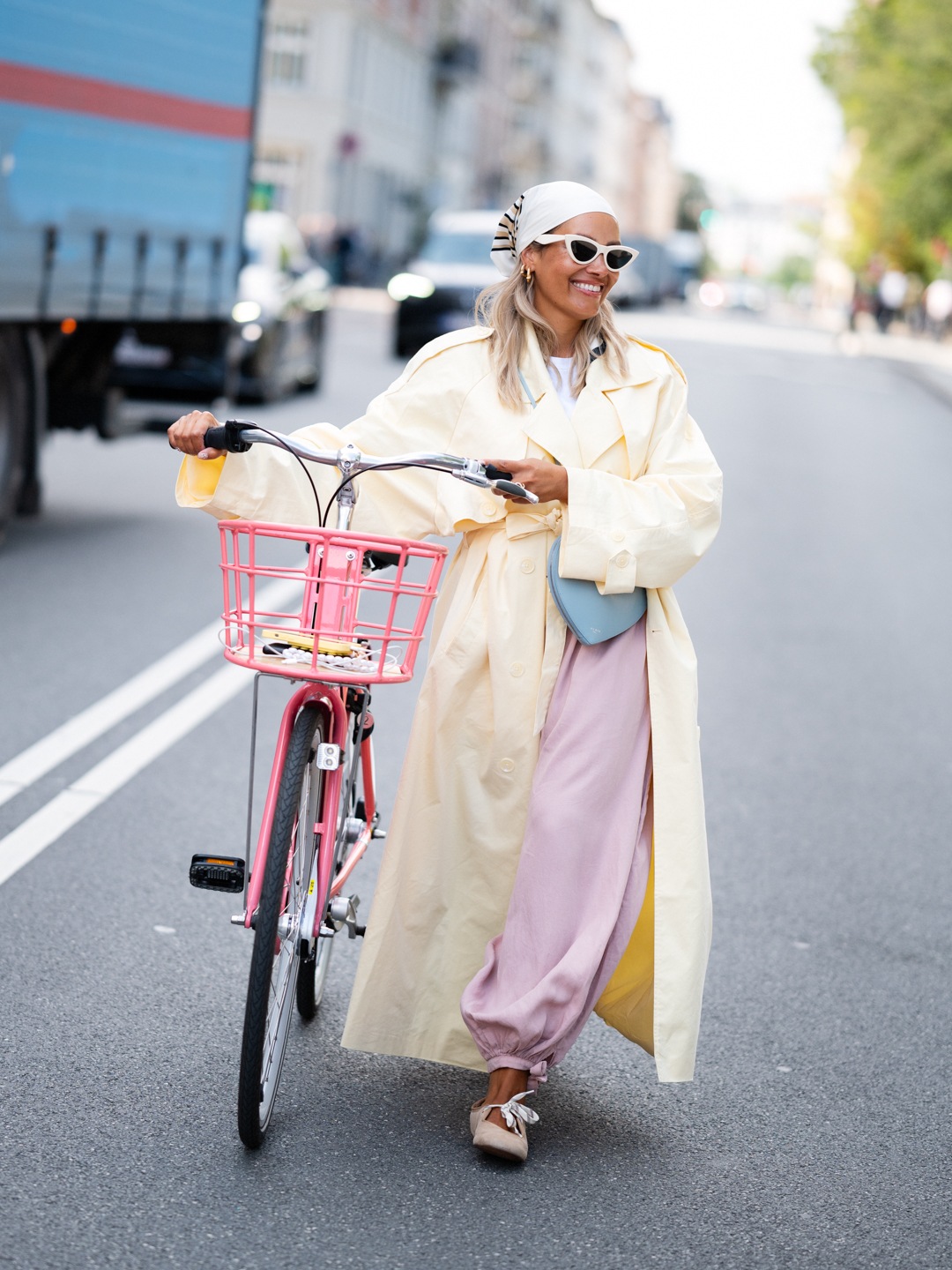 Nicole Reed smiling beside a pink bicycle in a pale yellow coat, pink trousers and white cat-eye sunglasses