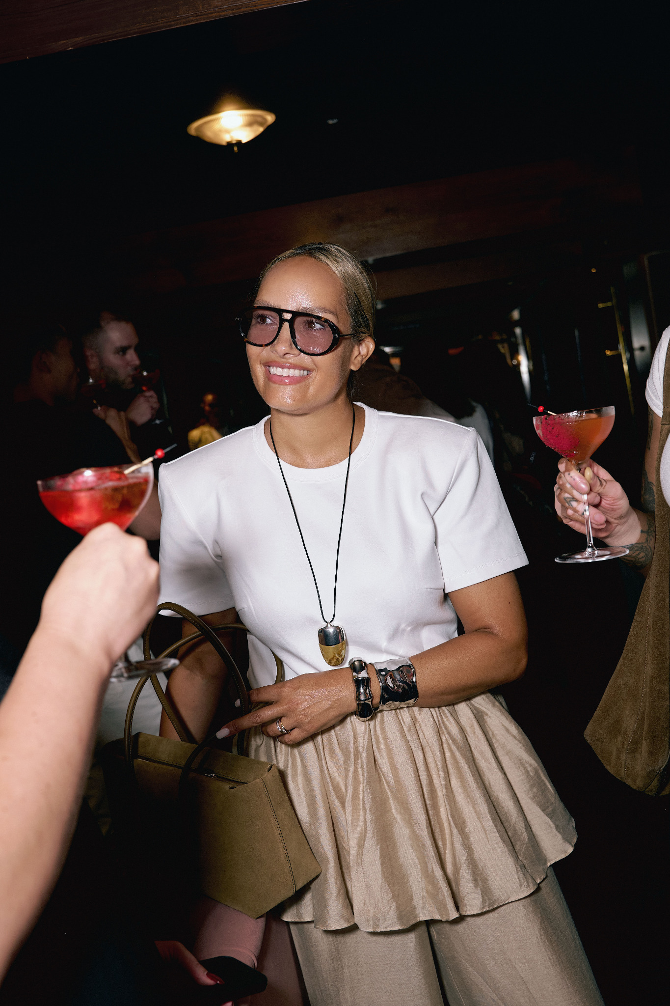 Nicole smiling among friends in oversized glasses, a white top and a beige skirt, with a sculptural pendant and silver cuff