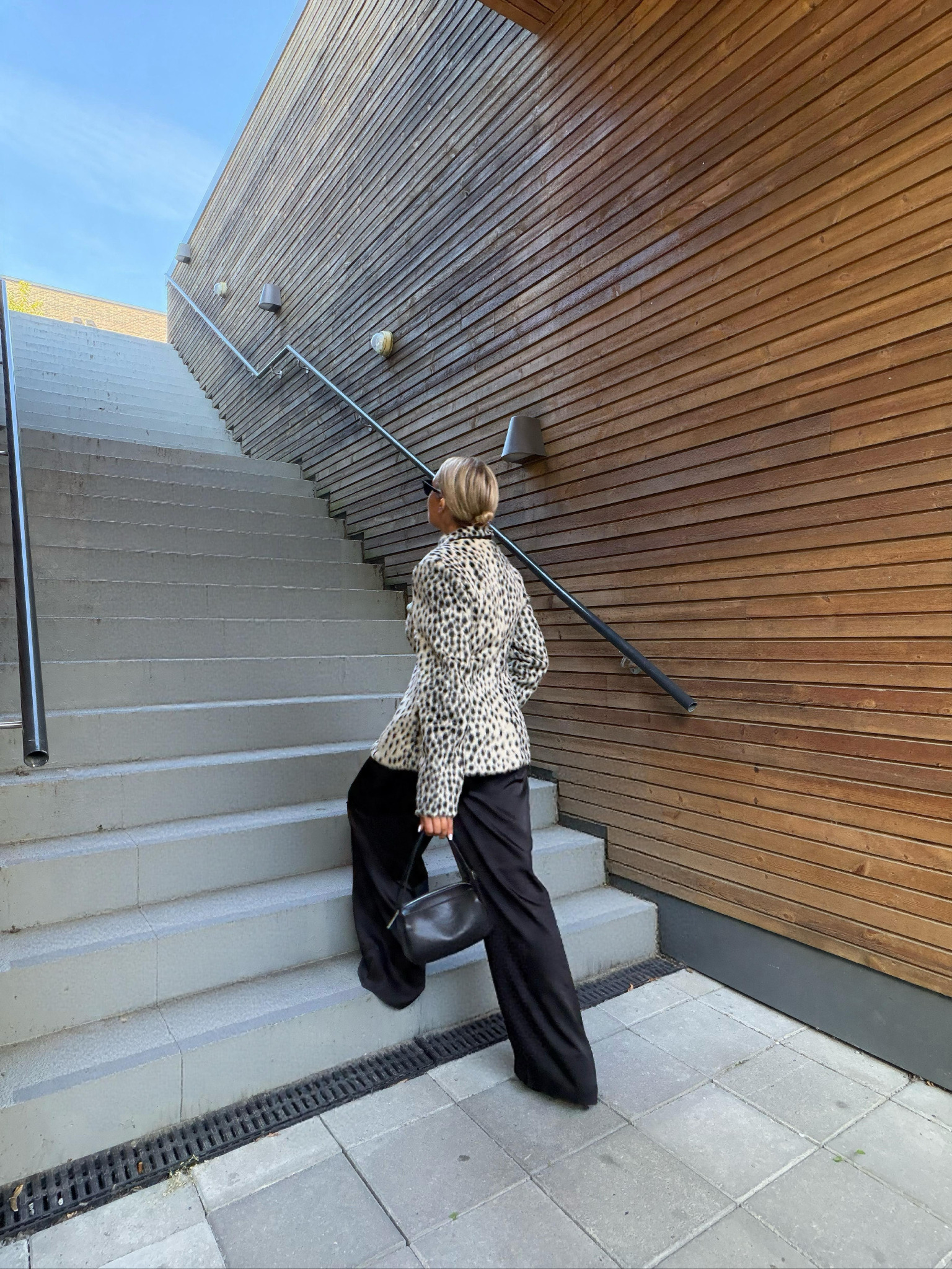 Nicole walking up outdoor stairs in a leopard-print jacket and wide black trousers, carrying a black handbag